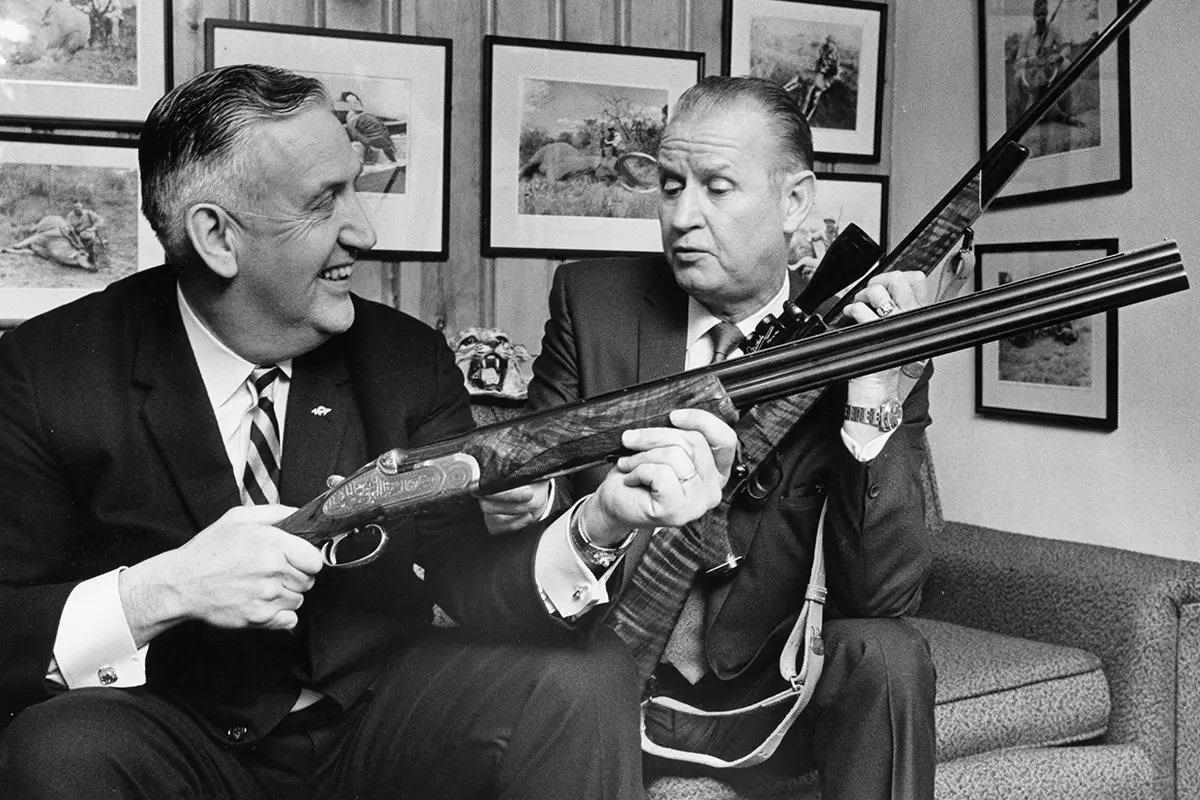 an old black and white book image with two men in suits sitting on a couch with old shotguns.